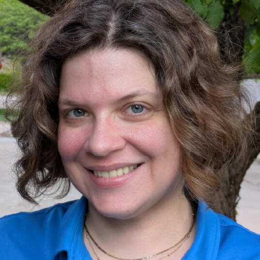 A woman with pale skin, brown short curly hair, and blue eyes. She is smiling and wears a bright teal polo shirt.